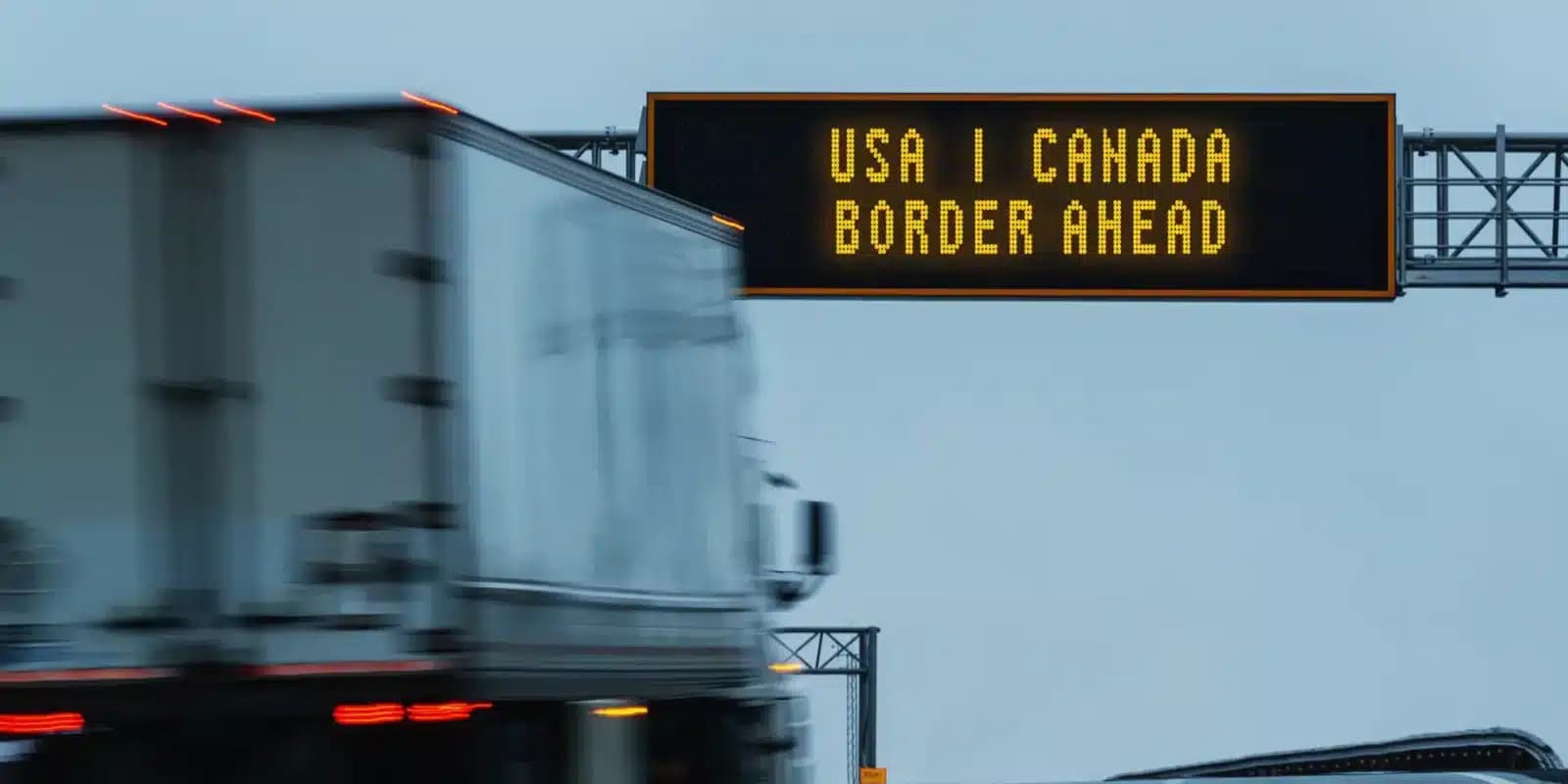 A tractor-trailer passing under an overhead sign reading USA Canada border ahead